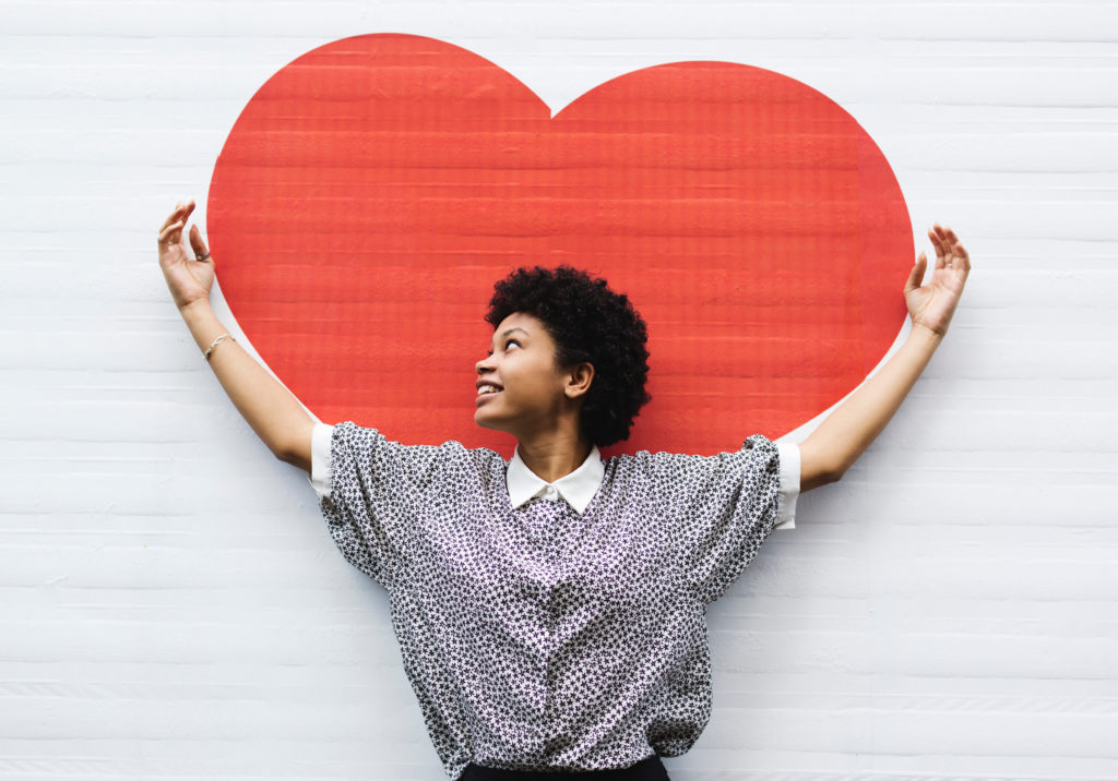Young woman smiling while standing in front of a big red heart mural with her hands up