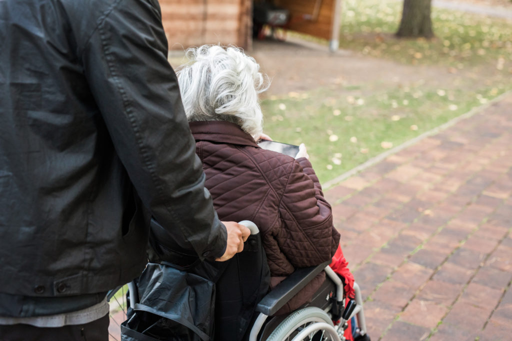 Man wearing black clothes pushes an older person in a wheelchair taking her for a walk in the park