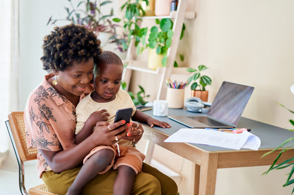A woman working in her home office takes a break to spend time with her child. They are looking online on a smartphone together