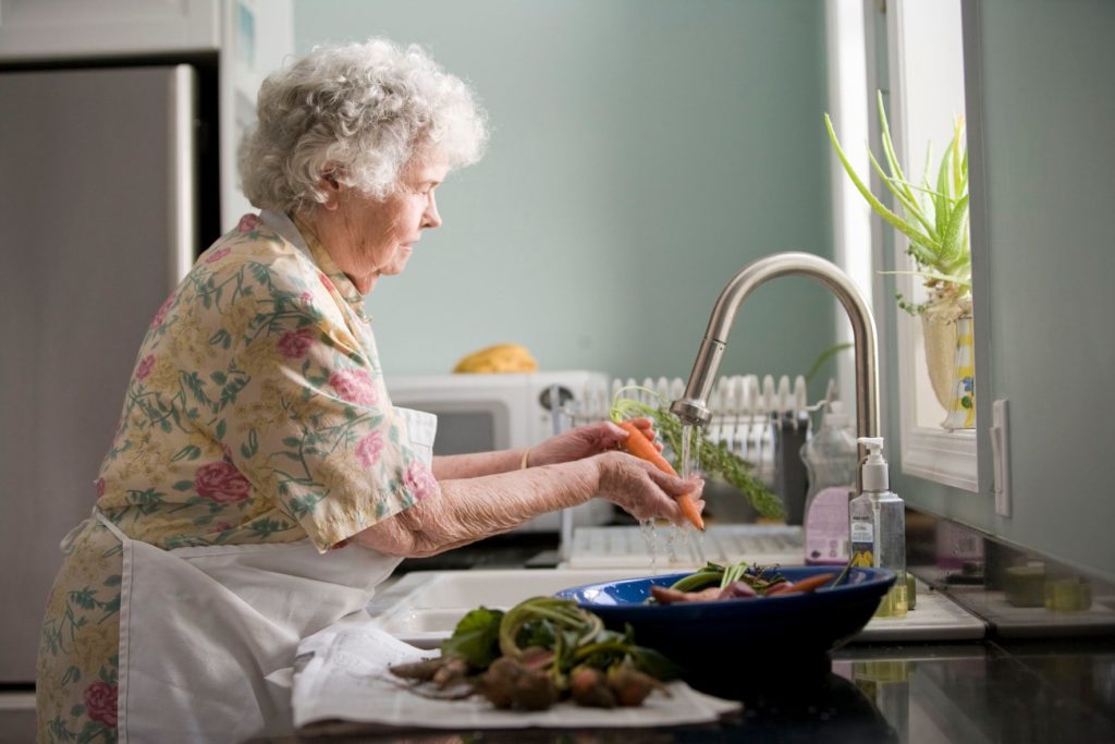 An elderly woman washing vegetables in her kitchen sink and getting ready to cook.
