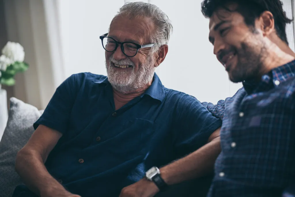 Elderly man and son sitting on a couch, smiling.