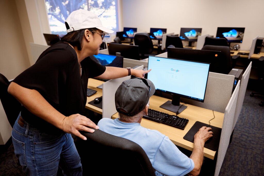 Woman showing a web browser to a senior man in a room filled with computer stations.