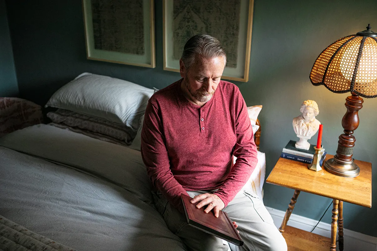 Man sitting on a bed in a dimly lit bedroom holding a book.